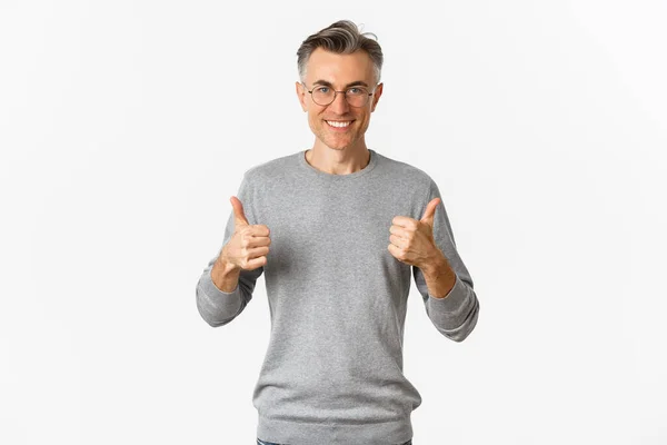 Imagem de homem de meia-idade confiante e satisfeito, sorrindo satisfeito, mostrando polegares para cima, vestindo camisola cinza e óculos, de pé sobre fundo branco — Fotografia de Stock