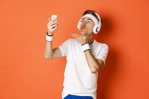Portrait of attractive middle-aged man in gym uniform, wearing headphones, sending air kiss at mobile phone camera, taking selfie or having video call, standing over orange background