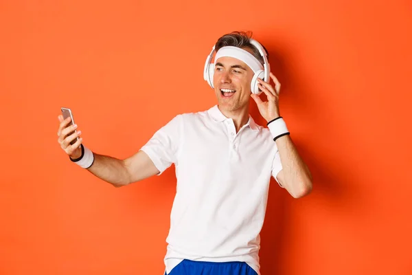 Porträt eines gut aussehenden männlichen Athleten mittleren Alters, der Turnuniform trägt, Musik über Kopfhörer hört und während des Trainings ein Selfie mit dem Handy macht, vor orangefarbenem Hintergrund steht — Stockfoto