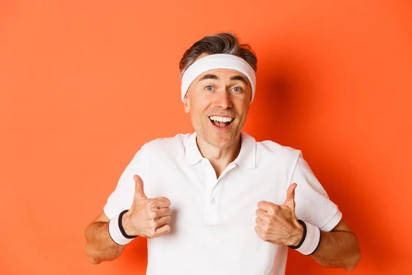 Close-up of cheerful middle-aged male athlete, wearing headband and t-shirt for workout, showing thumbs-up, approve something good, standing over orange background — Stock Photo, Image