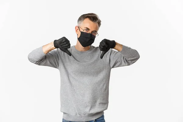 Fogalom a koronavírus, életmód és karantén. Portré szórakozás nélküli és szkeptikus középkorú férfi nem szeret valamit, visel orvosi maszk és kesztyű, mutatja hüvelykujj lefelé — Stock Fotó