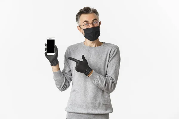 Concept of covid-19, social distancing and lifestyle. Image of attractive middle-aged guy in glasses, medical mask and gloves, explain something and pointing at mobile phone screen — Stock Photo, Image