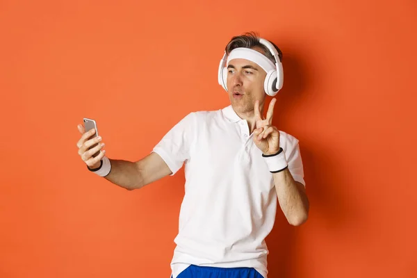 Portret atrakcyjnego sportowca w średnim wieku, robienie selfie podczas treningu, pokazywanie znaku pokoju, słuchanie muzyki w słuchawkach, stanie nad pomarańczowym tłem — Zdjęcie stockowe
