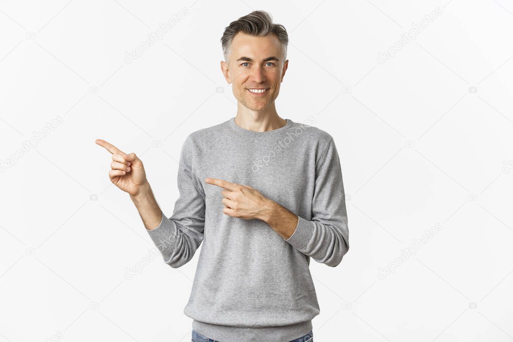 Image of handsome middle-aged man with grey short hairstyle, smiling pleased as pointing fingers left, showing logo on copy space, standing over white background.