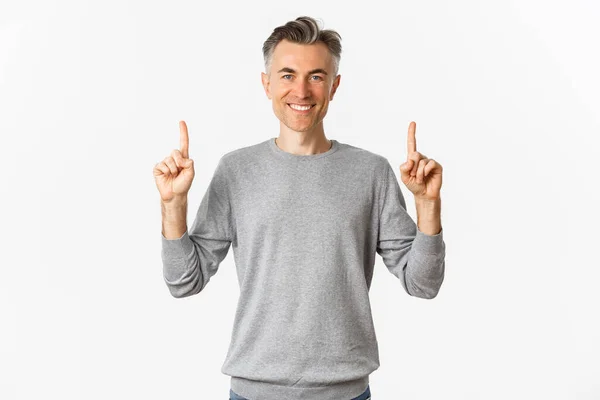 Портрет красивого успешного мужчины с серой короткой прической, улыбающийся приятно и указывая пальцами на логотип, показывая промо-предложение, стоя на белом фоне — стоковое фото