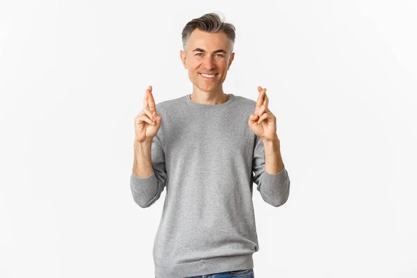 Изображение обнадеживающего мужчины средних лет, загадывающего желание, скрещивающего пальцы и улыбающегося, ожидающего хороших новостей, стоящего на белом фоне — стоковое фото
