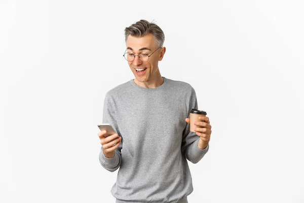 Image d'un bel homme d'âge moyen en pull gris et lunettes, buvant du café et regardant avec bonheur l'écran du smartphone, lisant quelque chose en ligne, debout sur fond blanc — Photo