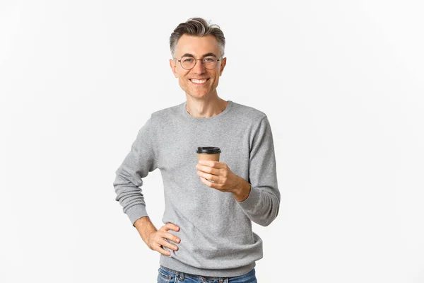 Bild eines selbstbewussten und gut aussehenden Mannes mittleren Alters, der Brille und grauen Pullover trägt, glücklich lächelt und Kaffee aus der Imbisstasse trinkt, zufrieden vor weißem Hintergrund steht — Stockfoto