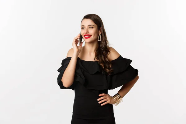 Успішна молода жінка в чорній сукні, червона помада і макіяж, розмовляє на мобільному телефоні і посміхається, стоїть над білим тлом — стокове фото