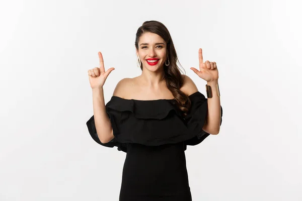 Moda y belleza. Encantadora mujer con labios rojos, vestido negro, sonriendo feliz y señalando con los dedos hacia arriba, mostrando el logotipo, fondo blanco —  Fotos de Stock