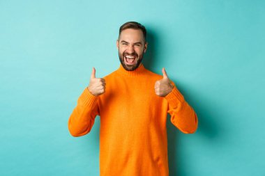 Süper iyi. Memnuniyet duyan mutlu adam sana katılıyor, mükemmel bir iş çıkarıyor, memnun görünüyor, turuncu süveter içinde açık mavi arka planda duruyor.