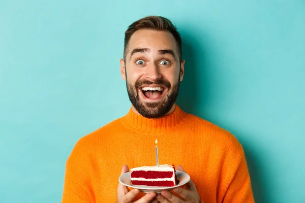 Close-up de homem adulto feliz comemorando aniversário, segurando bolo bday com vela e fazendo desejo, de pé contra fundo turquesa — Fotografia de Stock