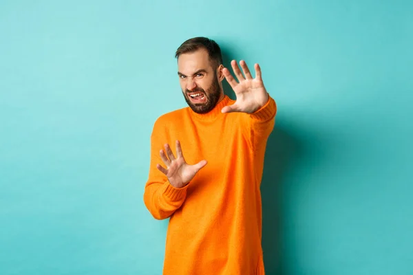 Un homme dégoûté grince et tend les mains pour se protéger, implorant miséricorde, regardant quelque chose avec aversion, debout sur un fond bleu clair — Photo