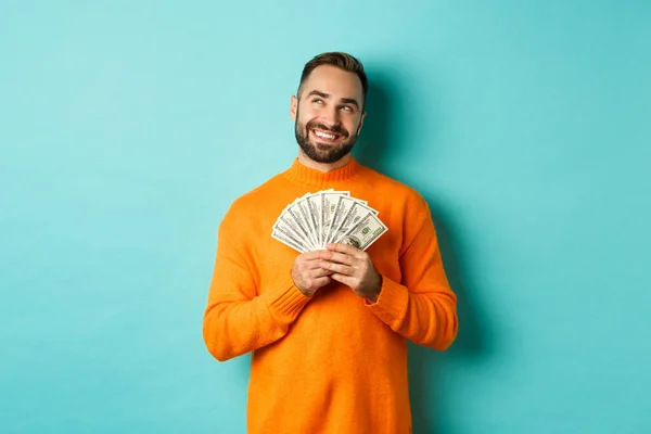 Hombre pensando en ir de compras, sosteniendo el dinero y sonriendo satisfecho, mirando a la esquina superior izquierda, de pie sobre fondo azul claro — Foto de Stock