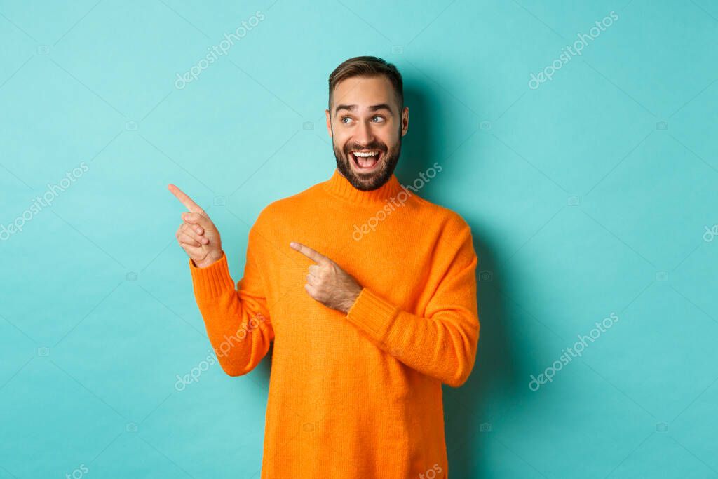 Handsome man looking happy and pointing fingers at upper left corner logo banner, standing in orange sweater on studio background.
