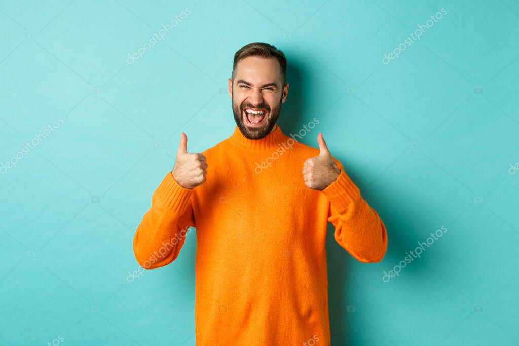Super good. Satisfied happy man showing thumbs up, agree with you, praise excellent work, looking pleased, standing in orange sweater against light blue background.