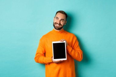 Teknoloji. Yakışıklı yetişkin adam dijital tablet ekranı gösteriyor, gülümsüyor ve sol üst köşeye bakıyor düşünceli, hayalperest ya da hayalperest, mavi arka plan