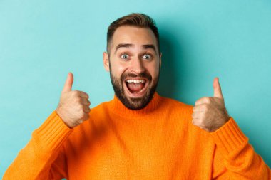 Heyecanlı yakışıklı adamın yakın plan çekimi başparmak kaldırması, ürün önermesi, harika kaliteyi övmesi, açık mavi arka planda durması.