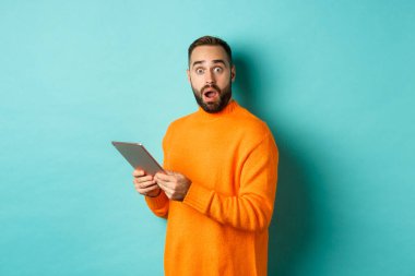 Turuncu süveter giyen, dijital tablet kullanan, açık mavi arka planda duran erkek manken görüntüsü