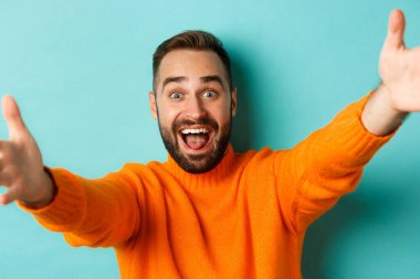 Yakışıklı, mutlu bir adamın yakın çekimi. Ellerini öne uzatıyor, kucaklaşmak için kollarını uzatıyor, turkuaz arka planda duruyor.