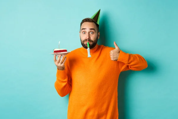 Happy birthday guy celebrating, wearing party hat, blowing wistle and holding bday cake and showing thumb-up in approval, standing over turquoise background — Stock Photo, Image