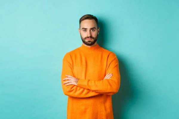 Jovem confiante olhando determinado, braços cruzados no peito, vestindo suéter laranja inverno, de pé contra fundo estúdio turquesa — Fotografia de Stock
