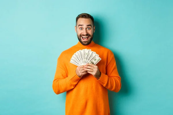 Imagen de tipo barbudo excitado sosteniendo dinero y regocijándose, ganando premio en efectivo, de pie sobre fondo azul claro — Foto de Stock