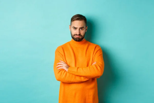 Stupide gars triste avec barbe, boudant et regardant offensé, bras croisés sur la poitrine et fronçant les sourcils bouleversés, debout sur fond bleu clair — Photo