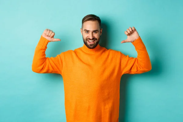 Handsome confident man pointing at himself, looking self-assured, standing in orange sweater against light blue background — Stock Photo, Image