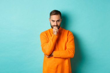 Düşünceli genç adam karar veriyor, ciddi görünüyor ve düşünüyor, seçiyor, fotokopi alanı turkuaz arka planının yanında duruyor.