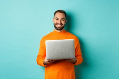 Laptopta çalışan ve gülümseyen, açık mavi arka planda turuncu süveter giyen yakışıklı bir serbest yazar.