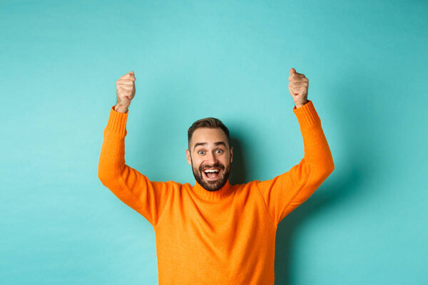 Талия счастливого человека, поднимающего руки, как будто держащего знак, показывающего логотип или рекламный баннер, улыбающегося взволнованного, стоящего в оранжевом свитере на бирюзовом фоне