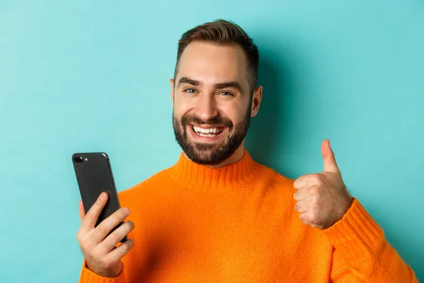 Primer plano del hombre barbudo sosteniendo el teléfono inteligente, pulgares hacia arriba, recomendando la aplicación móvil, de pie satisfecho sobre fondo turquesa — Foto de Stock