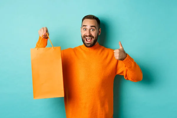 Hombre feliz sosteniendo bolsa de compras naranja y regocijándose de descuento y celebración, mostrando los pulgares hacia arriba y recomendando, de pie sobre fondo azul —  Fotos de Stock