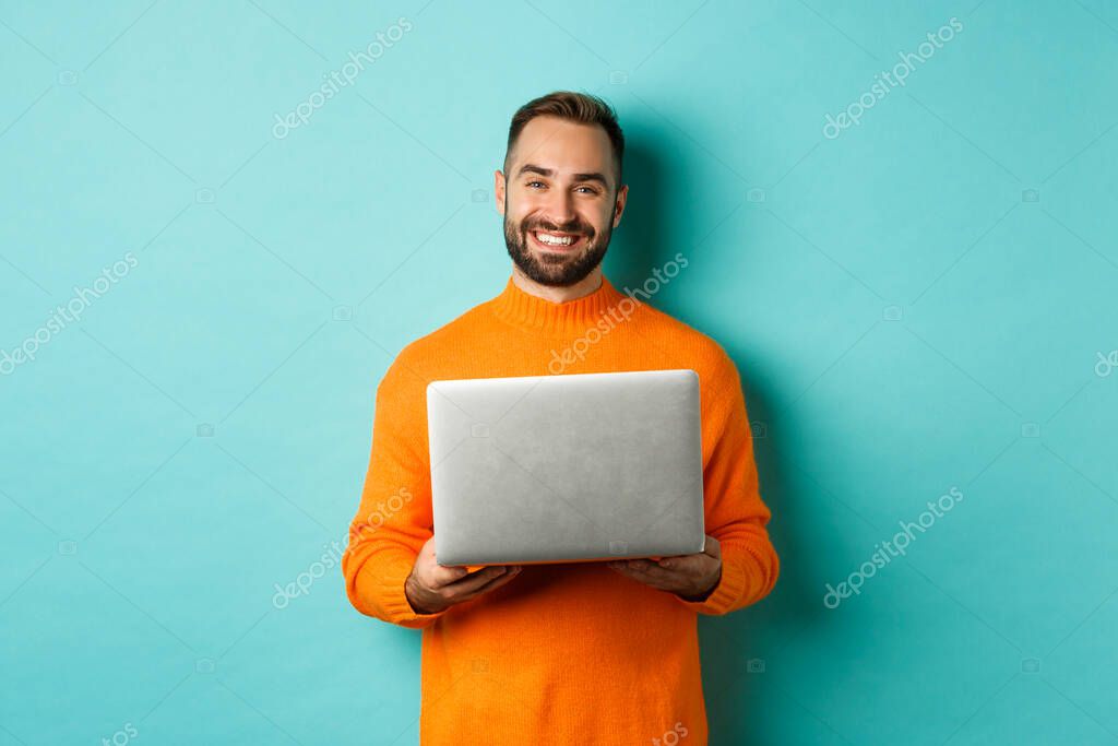 Handsome freelancer working on laptop and smiling, standing in orange sweater over light blue background.