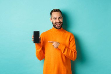 Turuncu kazaklı yakışıklı adam, cep telefonu ekranını işaret ediyor, akıllı telefon uygulamasını gösteriyor, açık mavi arka planda duruyor.