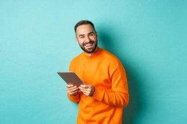 Yakışıklı sakallı adam dijital tablet kullanıyor, kameraya gülüyor, açık mavi arka planda mutlu duruyor.