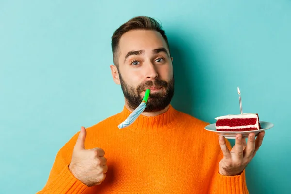 Primer plano del hombre adulto divertido celebrando su cumpleaños, sosteniendo pastel de bday con vela, soplando el silbato de la fiesta y mostrando el pulgar hacia arriba, de pie sobre el fondo azul claro — Foto de Stock