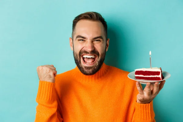 Close-up of happy adult man celebrating birthday, holding bday cake with candle and making wish, standing against turquoise background — Stock Photo, Image