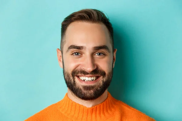 Headshot van knappe blanke man met baard lachend blij op camera, staande in oranje trui tegen turquoise achtergrond — Stockfoto