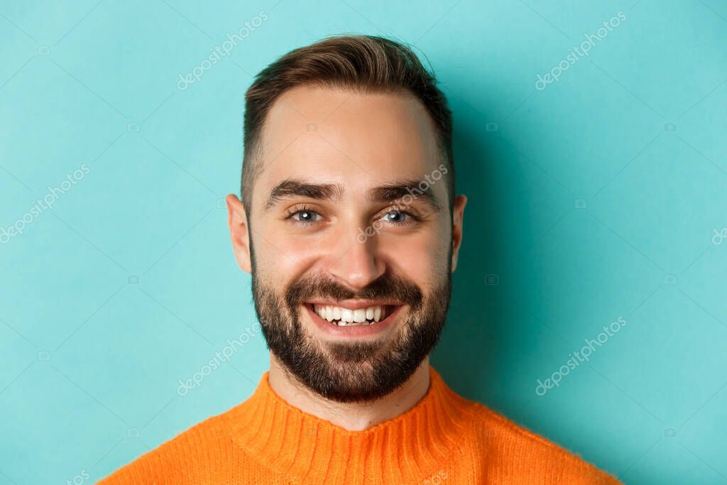 Headshot of handsome caucasian man with beard smiling happy at camera, standing in orange sweater against turquoise background.