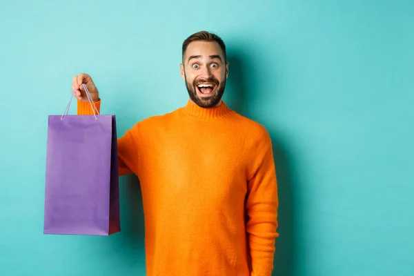 Hombre adulto excitado sosteniendo la bolsa de compras púrpura y sonriendo, yendo al centro comercial, de pie sobre fondo turquesa —  Fotos de Stock