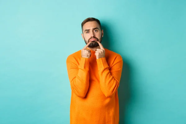 Imagem de homem barbudo sombrio, fazendo cara triste e franzina, de pé chateado em suéter laranja contra fundo turquesa — Fotografia de Stock