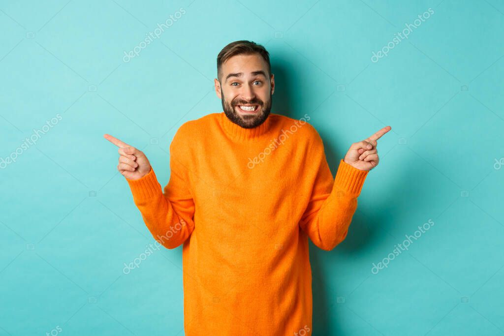 Confused handsome man shrugging indecisive, pointing fingers sideways, cant choose between two variants, standing against light blue background.