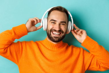 Kulaklıkla müzik dinleyen, turuncu süveter giymiş turkuaz arka planda duran yakışıklı modern adamın yakın çekimi.