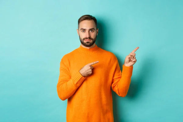 Handsome and serious bearded man in orange sweater pointing right, showing advertisement or logo, standing over turquoise background
