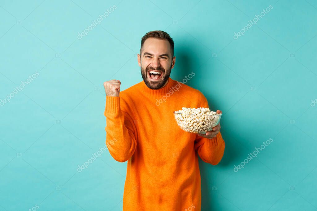 Cheerful handsome man in orange sweater, saying yes, cheering and rejoicing, eating popcorn and watching sports, making fist pump satisfied, standing over blue background.