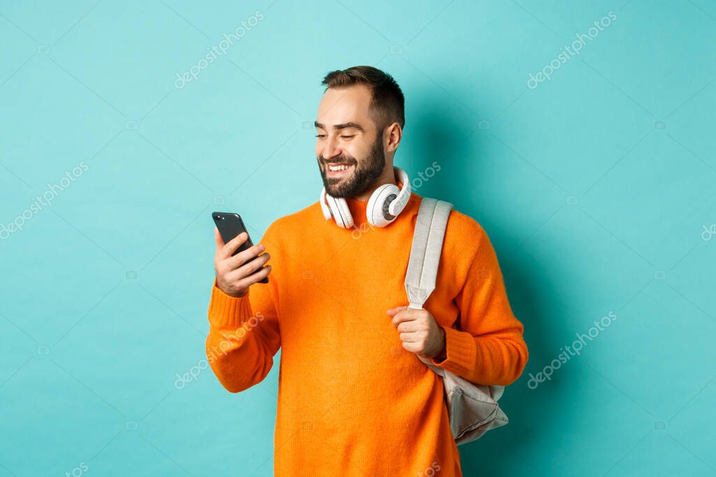 Caucasian man with headphones and backpack looking at phone, reading message and smiling, standing over turquoise background.