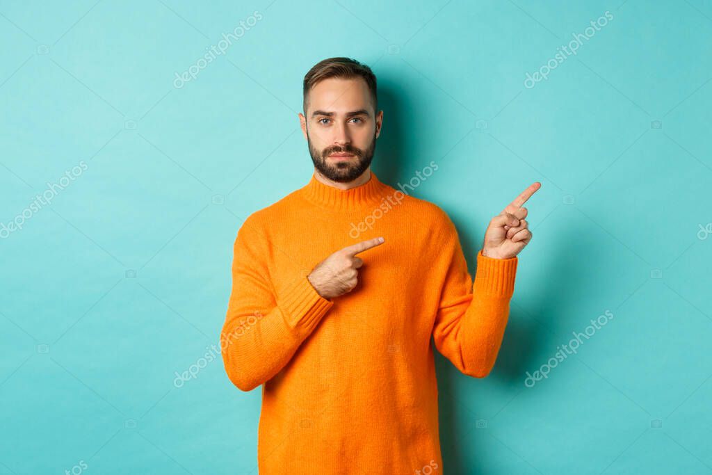 Handsome and serious bearded man in orange sweater pointing right, showing advertisement or logo, standing over turquoise background.