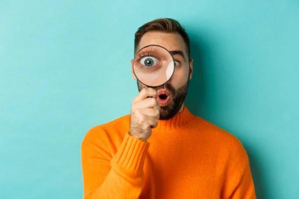 Funny man looking through magnifying glass, searching or investigating something, standing in orange sweater against turquoise background — Stock Photo, Image